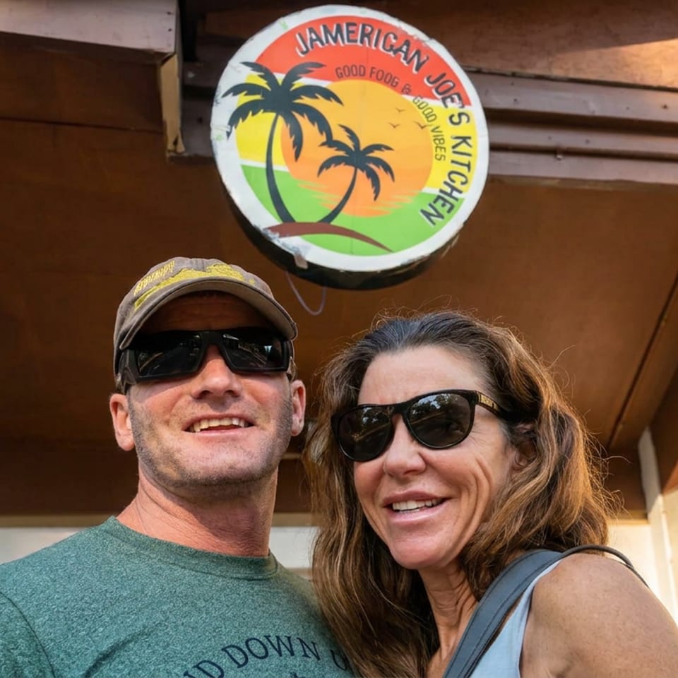 Smiling couple wearing sunglasses outside an American Joe’s Kitchen restaurant beneath a colorful palm-tree sign.